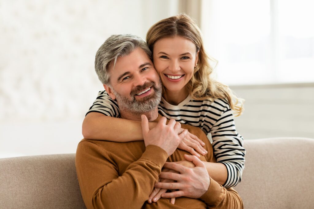 Implant-Supported Bridges Wauwatosa couple hugging each other. He is sitting on the couch and she is behind him.